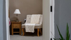 A cozy room with a beige armchair, a wooden nightstand with a lamp, and a small table. - Ceunant Bach - One Bedroom Cottage, Sleeps 2 (Bethesda)