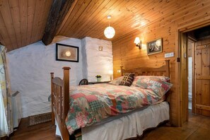Une chambre confortable avec un plafond en bois, un lit recouvert d’une courtepointe à motifs floraux, une table de chevet et une lampe fixée au mur.
