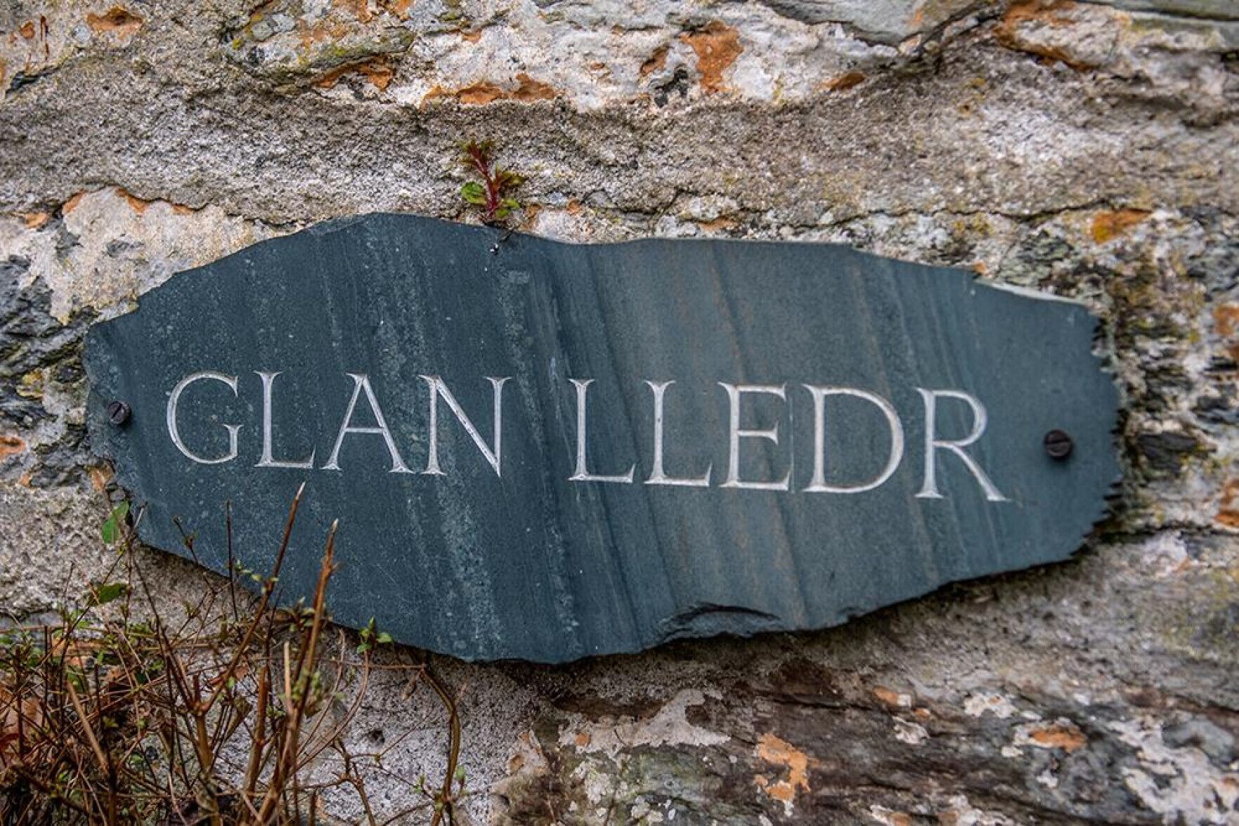 Ein dunkles Schiefer Schild mit der Aufschrift 'GLAN LLEDR', befestigt an einer Steinmauer.