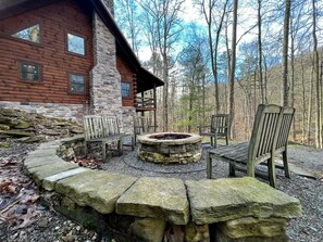 A rustic cabin with a stone fireplace, wooden benches, and a stone fire pit surrounded by a stone wall.