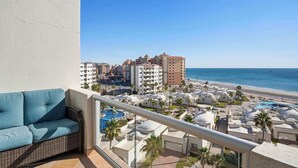 Un balcone con divano in vimini che si affaccia su una zona resort con piscina, palme e spiaggia.