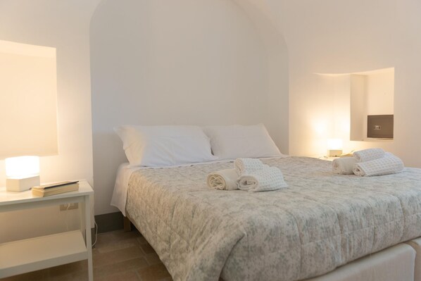 A neatly made bed with white linens and rolled towels, a bedside table with a lamp, and a wall-mounted TV.