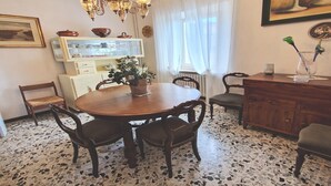 Una zona de menjador amb una taula rodona de fusta, sis cadirees, un penjó, un armari amb plats i una finestra amb cortines.