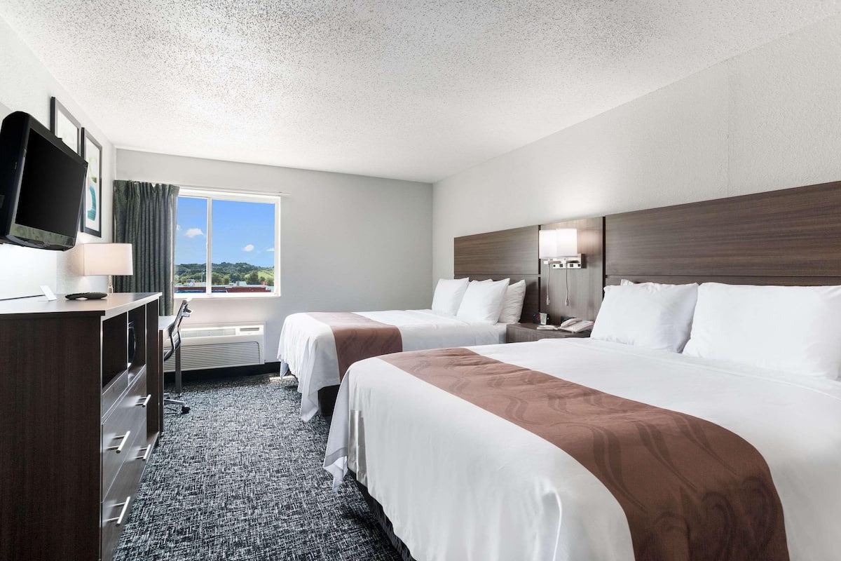A hotel room with two beds, a desk with a TV, and a window with a view of the outdoors.