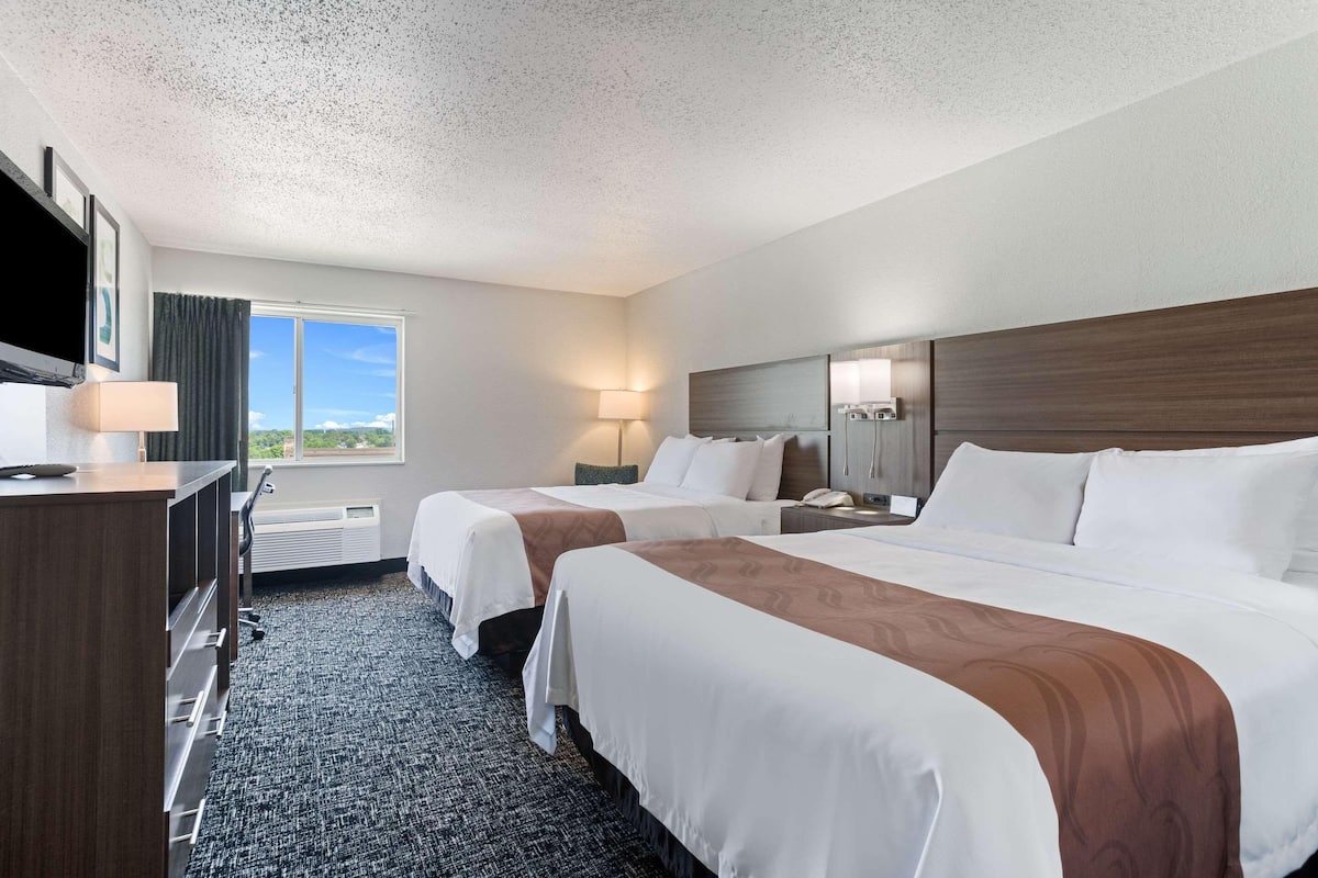 A hotel room with two beds, a desk, a TV, a window with a view, and a lamp.