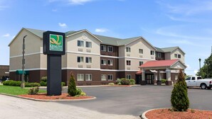 A hotel with a sign reading 'Quality Inn & Suites,' a parking lot, and a building with a red roof. - Quality Inn & Suites Ferdinand (Ferdinand)