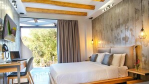A modern bedroom with a large bed, a desk, and a laptop. There is a window with curtains and a view of greenery outside. - Royal Hotel and Suites (Kassandra)