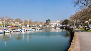Une rivière paisible où des bateaux sont amarrés le long des rives, un sentier piétonnier et des bâtiments en arrière-plan.