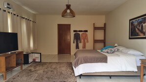 A hotel room with a bed, a TV, a wooden side table, a robe and towel rack, a painting, and a door. - Pousada Caminhos da Vila Chantal (Campos do Jordão)