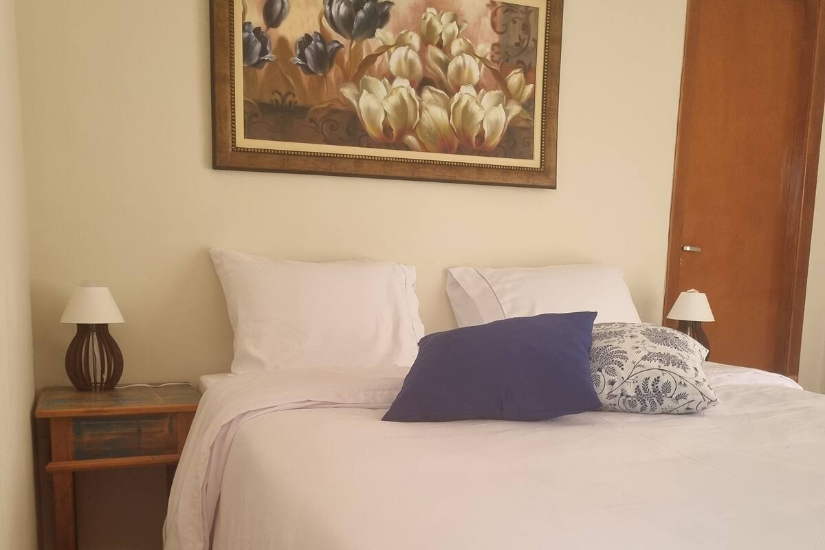 A neatly made bed with white and blue pillows, a wooden nightstand with a lamp, and a painting of flowers on the wall.