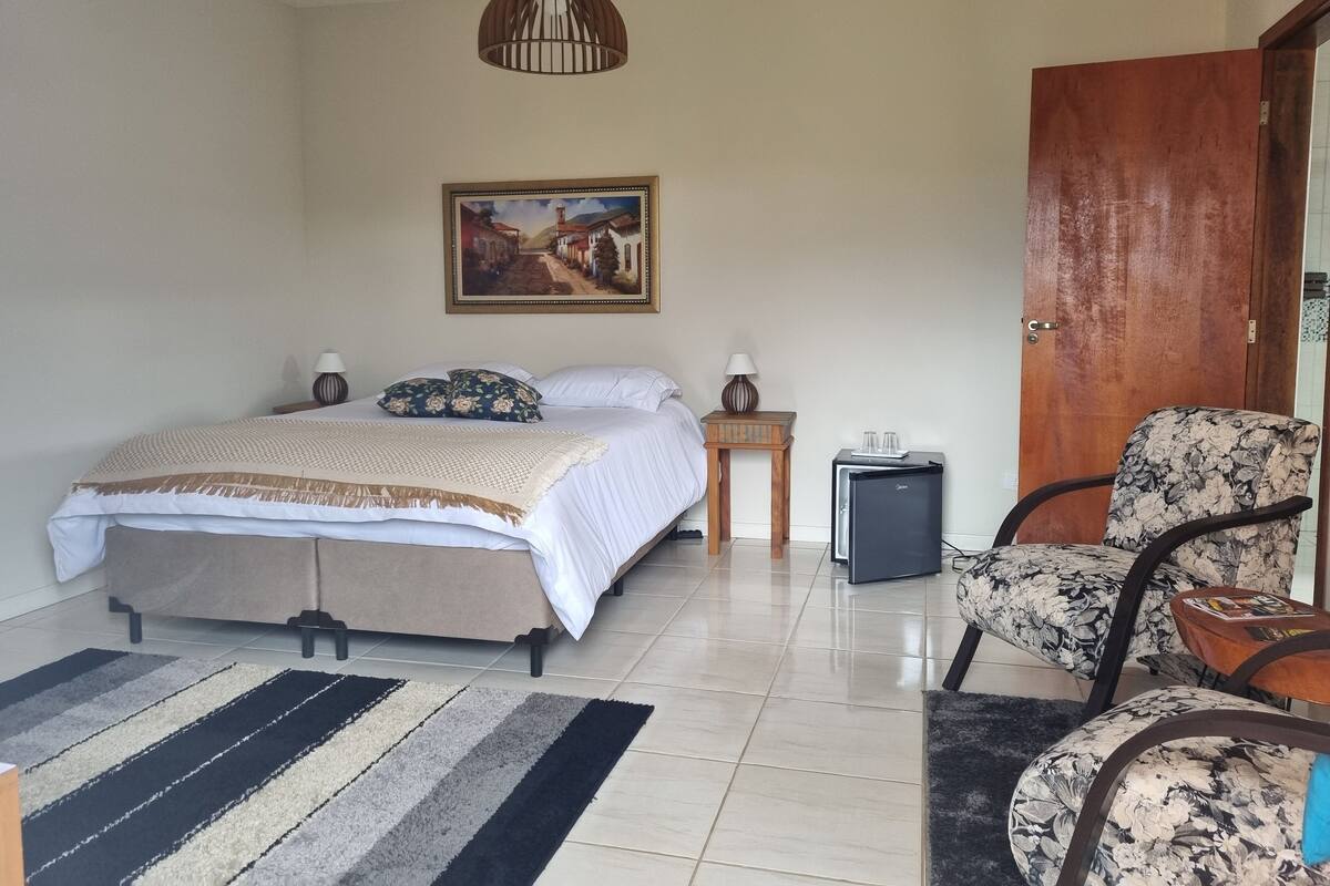 A bedroom with a bed, bedside tables, a refrigerator, a chair, a rug, a painting, and a wooden door.
