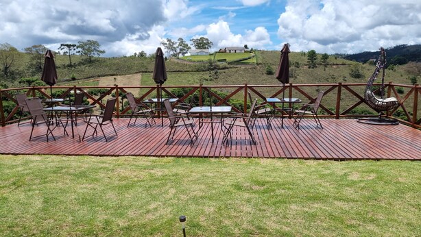 A wooden deck with outdoor seating, surrounded by a metal railing, overlooking a hilly landscape. - Pousada Caminhos da Vila Chantal (Campos do Jordão)
