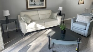 A modern living room with a white leather sofa, a glass coffee table, and a grey armchair. There are two side tables with lamps, and a framed picture of a beach scene on the wall. - -Near Disney-  condo in gated Bahama Bay Resort (Davenport)