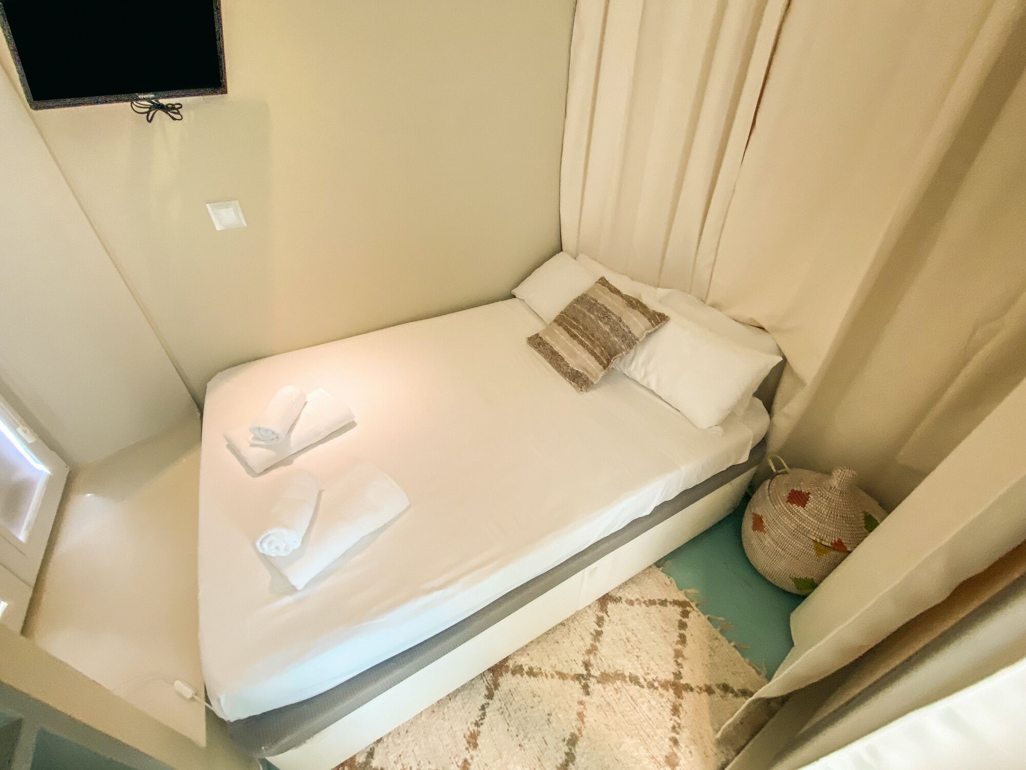 A small, single-bed room with a mattress, pillows, a blanket, a bedside table with a lamp, and a wall-mounted TV.