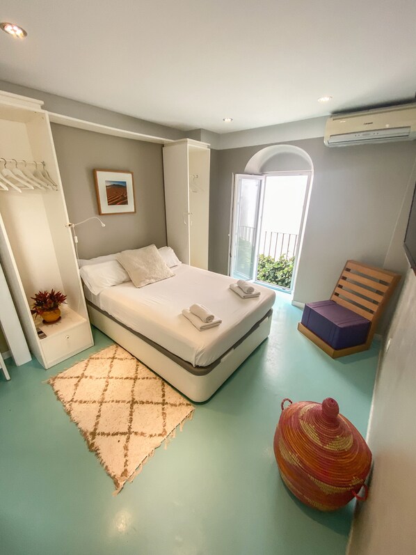 Un dormitorio con cama, mesita de noche con lámpara, una silla, una pequeña mesa con planta y ventana con vista a vegetación.