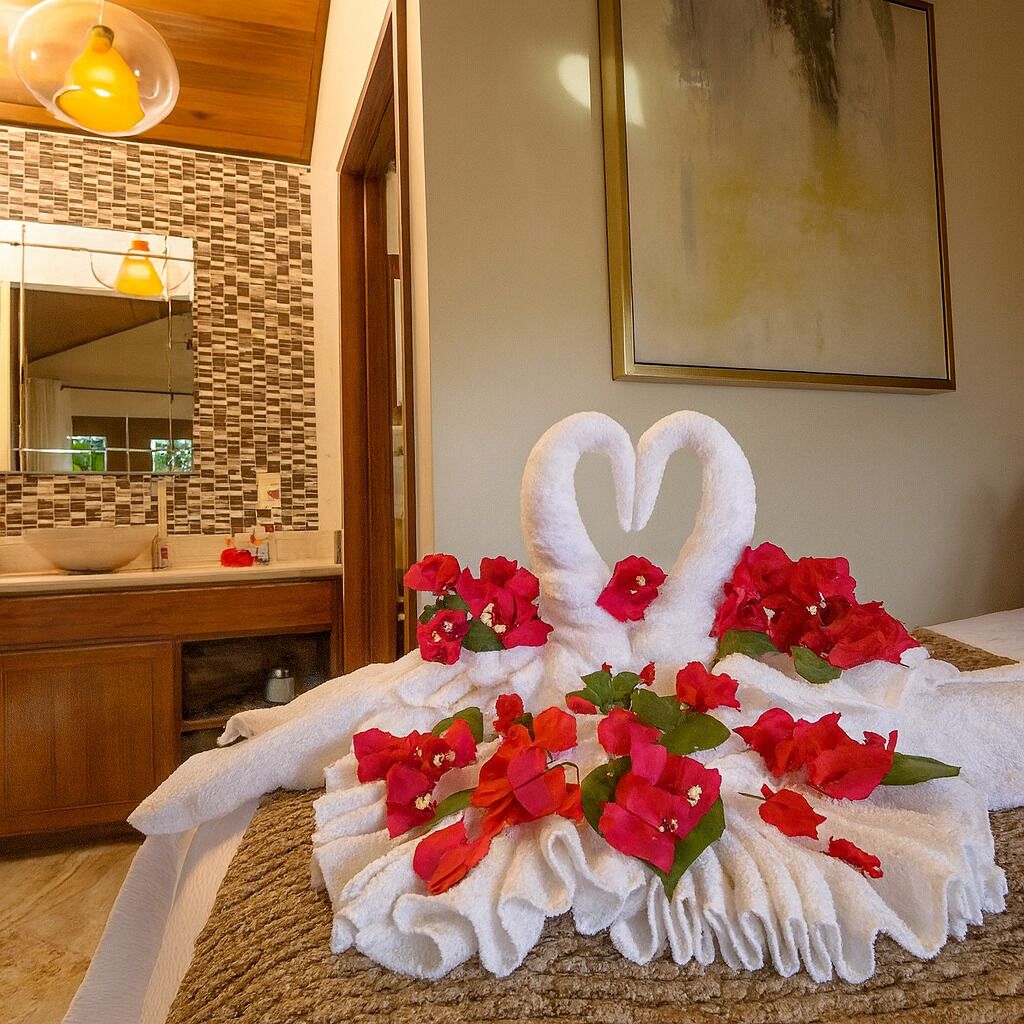 Una cama impecablemente arreglada con una toalla dispuesta en forma de cisne, decorada con flores rojas, en una habitación de hotel con suelo de madera, un salpicadero de azulejos y un espejo grande.
