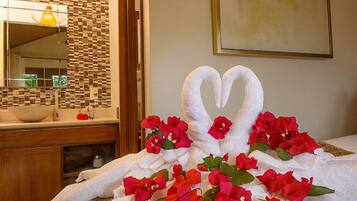 Una cama impecablemente arreglada con una toalla dispuesta en forma de cisne, decorada con flores rojas, en una habitación de hotel con suelo de madera, un salpicadero de azulejos y un espejo grande.