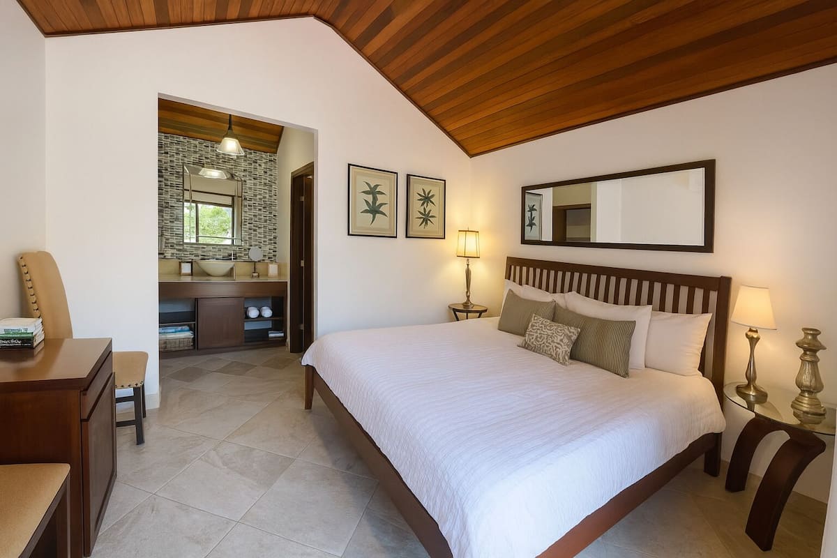 A bedroom with a large bed, bedside lamps, a wooden headboard, a desk with a chair, a vanity with a mirror, and framed artwork on the wall.