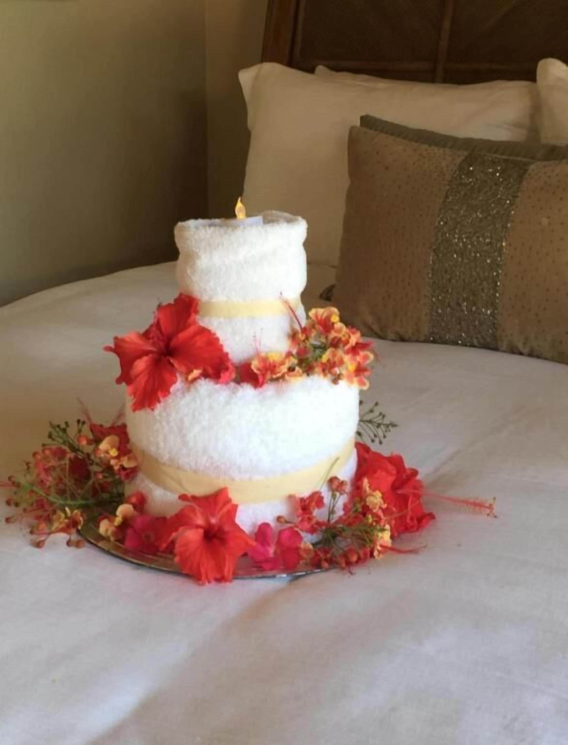 A three-tiered cake with white frosting and red flowers, placed on a bed with white bedding and a gold pillow in the background.