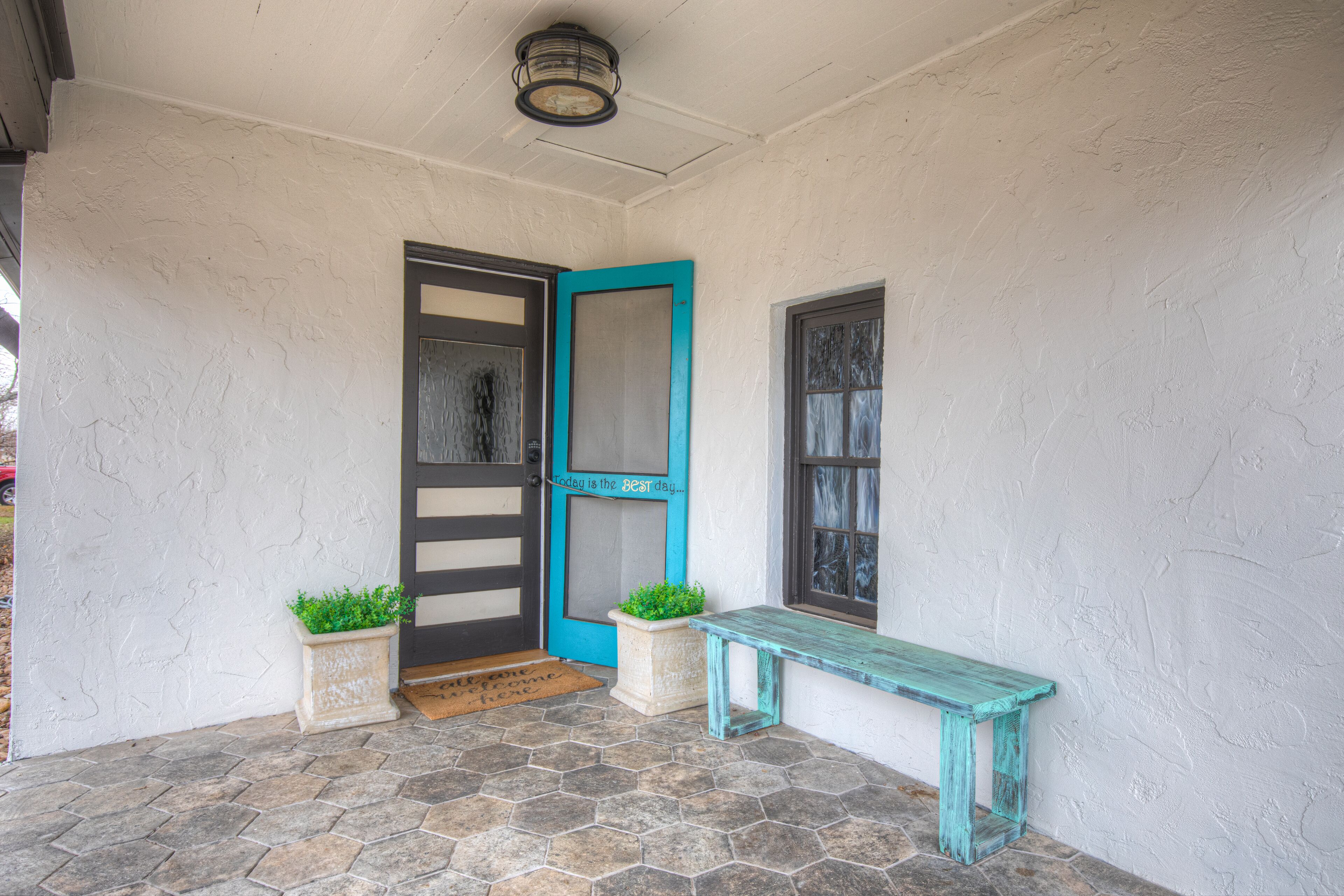 Un porche abrité, doté d’une porte, d’un banc et de deux jardinières ornées de plantes vertes.