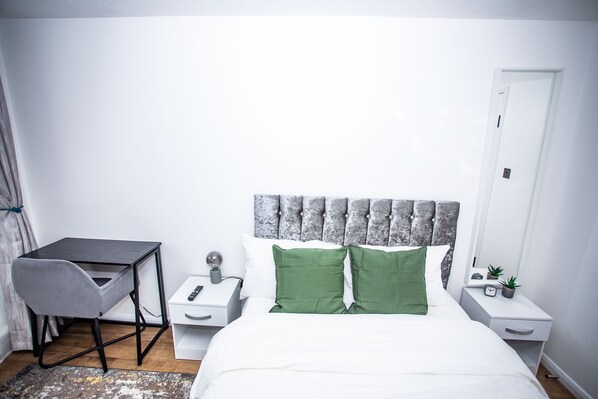 Un dormitorio con cama, escritorio, silla, mesita de noche y espejo, además de una puerta.