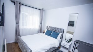 Un dormitorio moderno con una cama grande, una mesita de noche, una silla, un espejo y una ventana con cortinas.