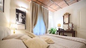 A hotel room with a large bed, a desk with a chair, a lamp, a mirror, and a window with curtains. - Corte Paolina - charming courtyard inside Lucca (Lucca)