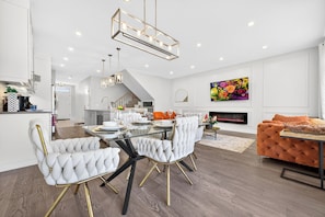 A modern kitchen with white cabinetry, a dining area with a glass table, and a living room with a fireplace.