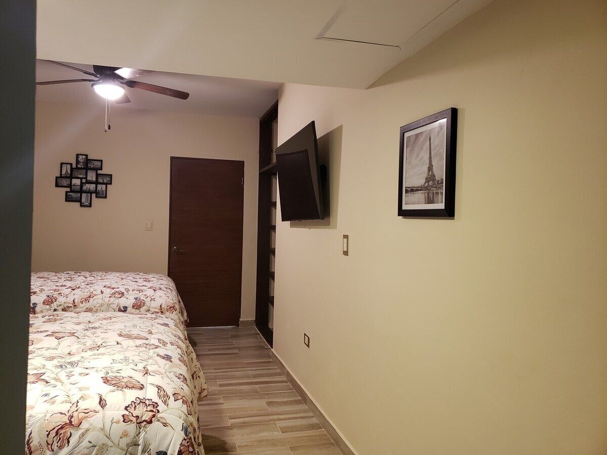 A bedroom with a bed, a ceiling fan, a TV mounted on the wall, and a door leading to another room.