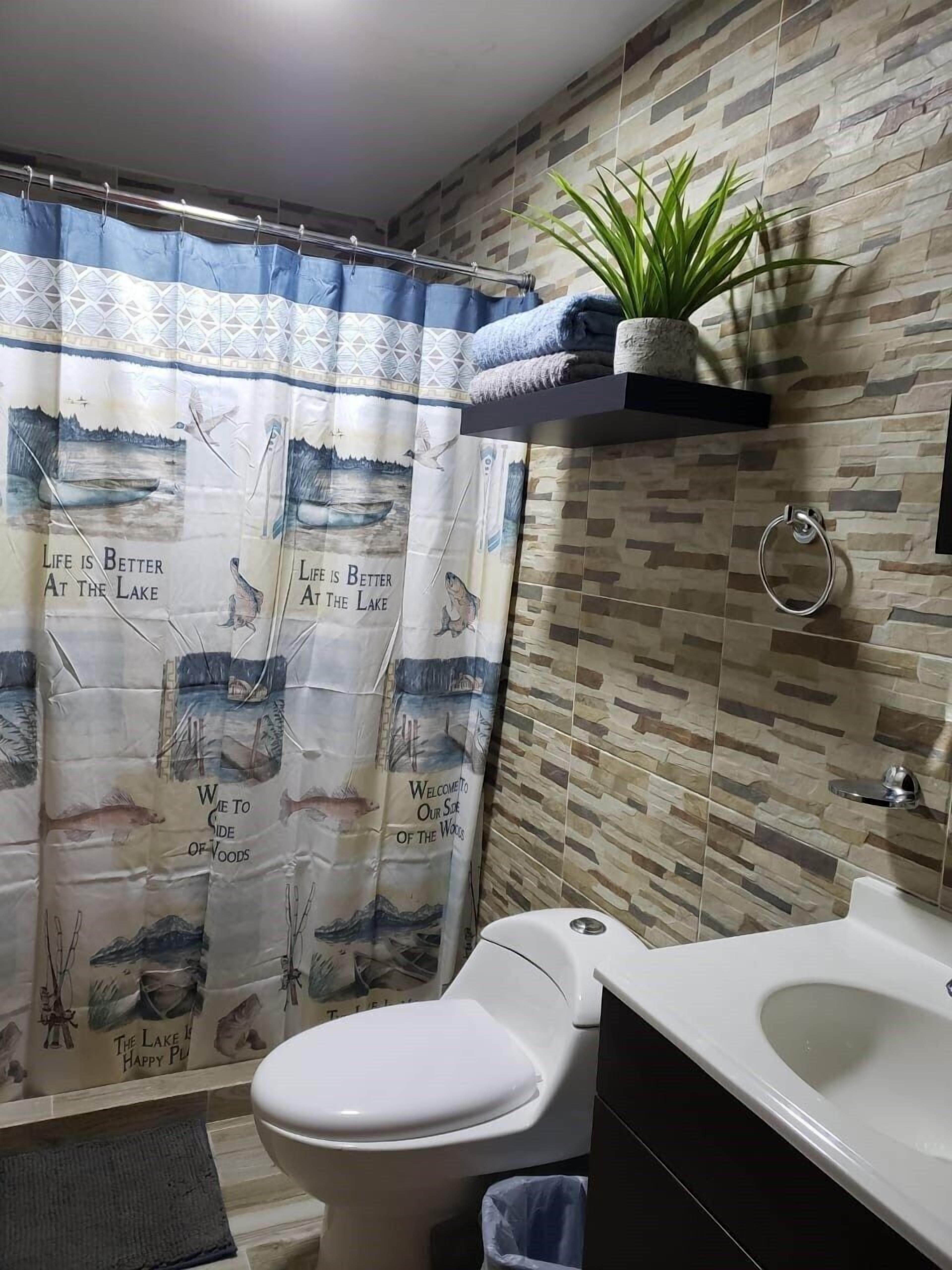 A bathroom with a shower curtain featuring a lake and wildlife theme, a toilet, a sink, and a small shelf with a plant.