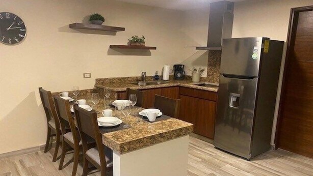 Cozinha compacta com ilha central, cadeiras de madeira, geladeira de aço inoxidável e um relógio na parede.