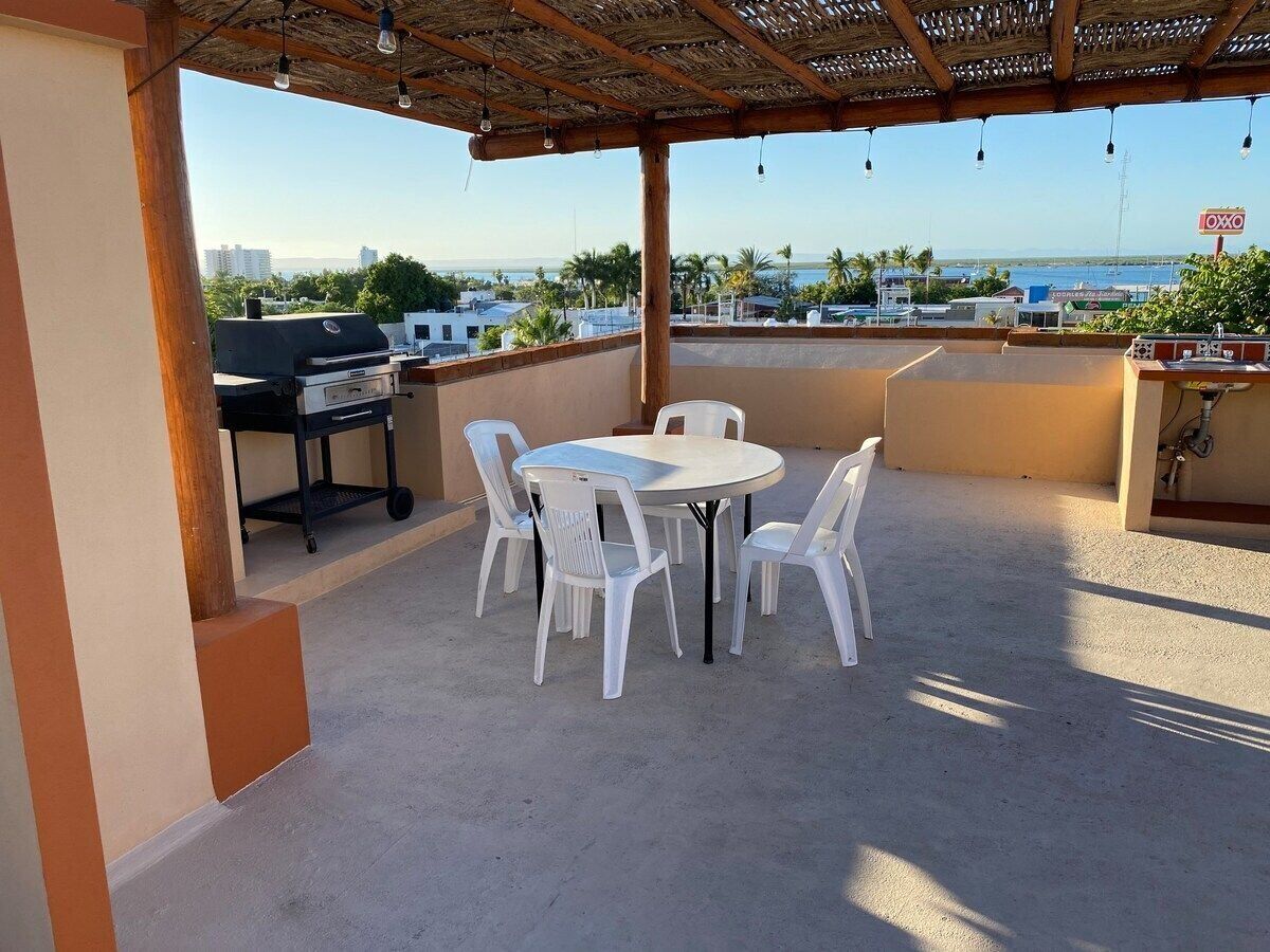 A covered outdoor patio with a round table and chairs, a grill, and a view of the sea and buildings in the background.