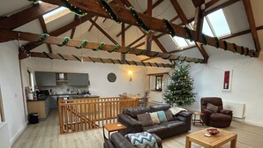A spacious living area with a high ceiling, wooden beams, a cozy brown leather sofa, a wooden coffee table, and a small wooden dining table. A Christmas tree with lights is visible in the background. - Tawny Cottage pet friendly, barn conversion, quiet farm location, nr Dartmouth. (Dartmouth)