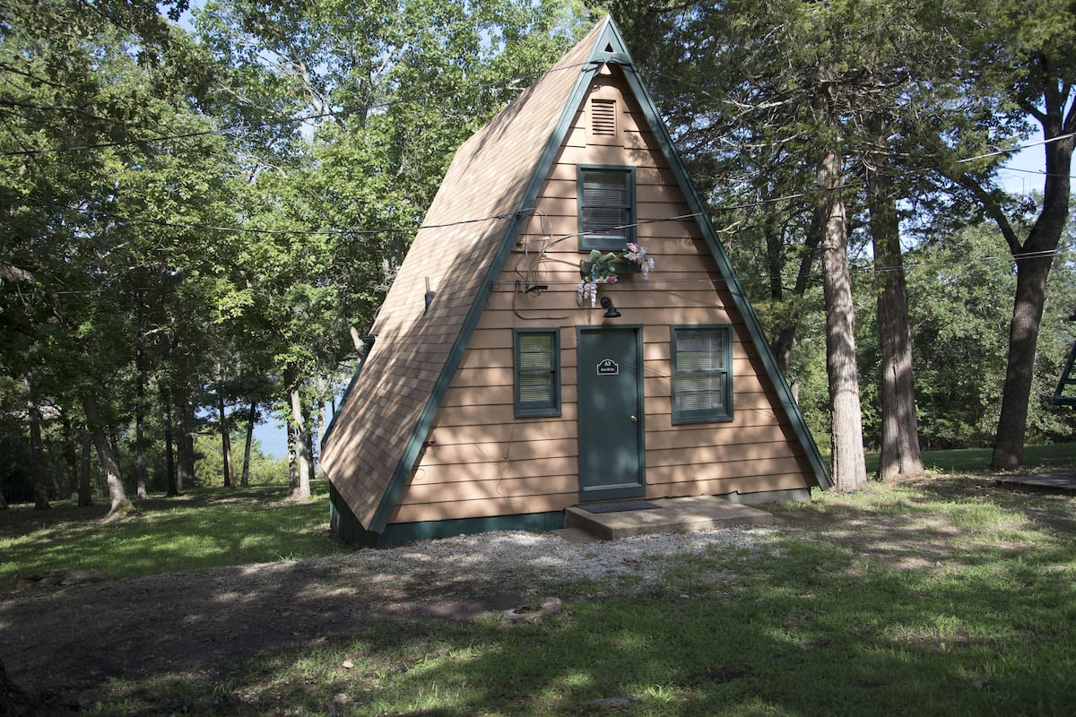 Uma cabana pequena e triangular, com porta e janelas verdes, rodeada por árvores.