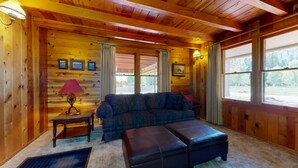 A cozy cabin interior with a wooden floor, a sofa, an ottoman, a lamp, and a window with curtains. - Spacious and Lovely Mountain Retreat (Bayfield)