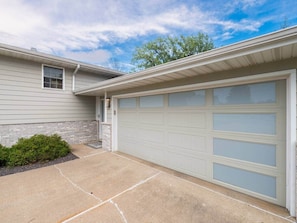 A residential garage with a light-colored exterior, a concrete driveway, and a small bush to the left.