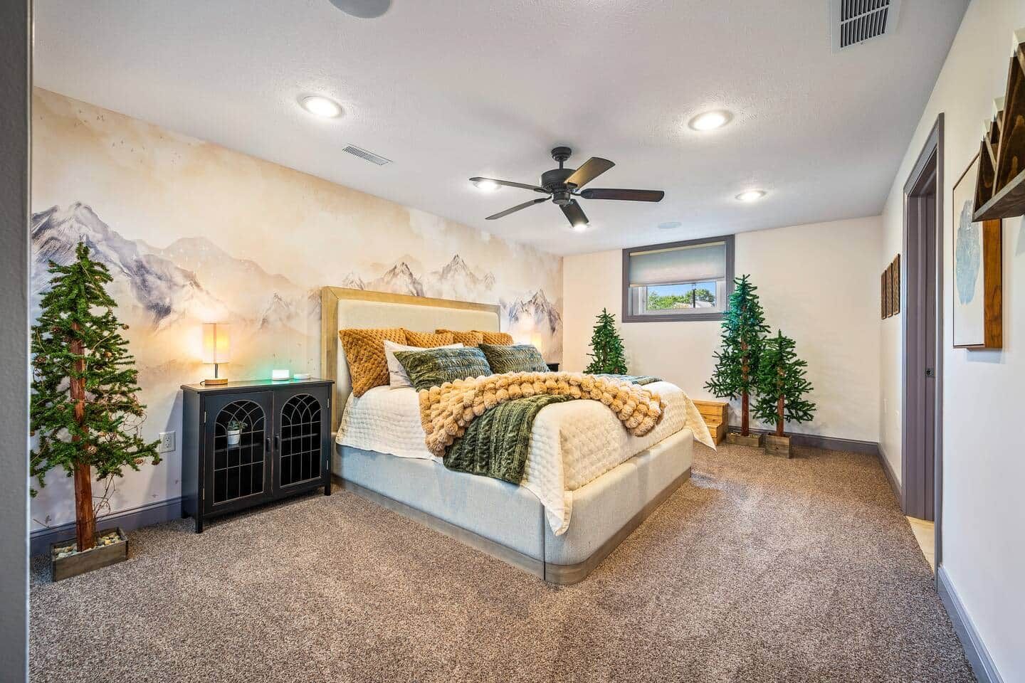 A bedroom with a bed, a ceiling fan, a nightstand, and two artificial trees.