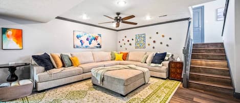 A living room with a sectional sofa, a ceiling fan, a world map artwork, a patterned rug, and a wooden staircase.