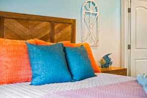 A bed with colorful pillows, a wooden headboard, a decorative wall piece, and a small blue fish figurine on a nightstand.