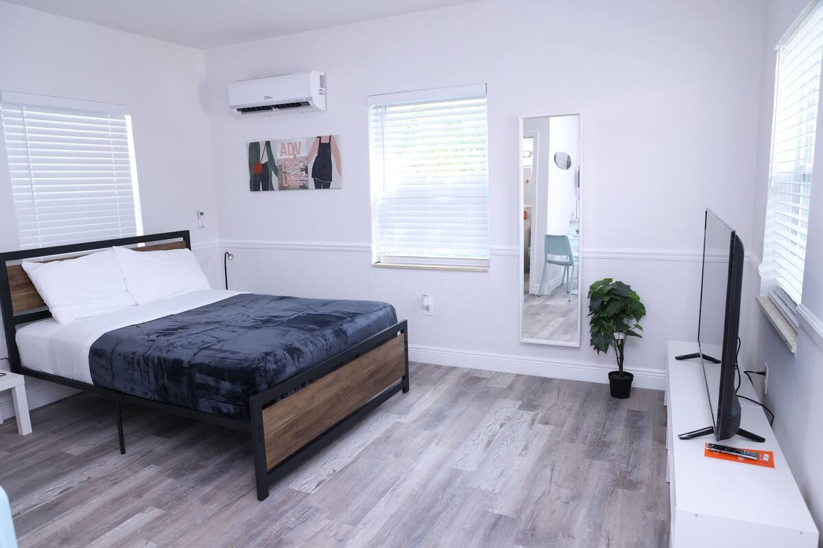 A bedroom with a bed, a TV, a potted plant, and a window with blinds.