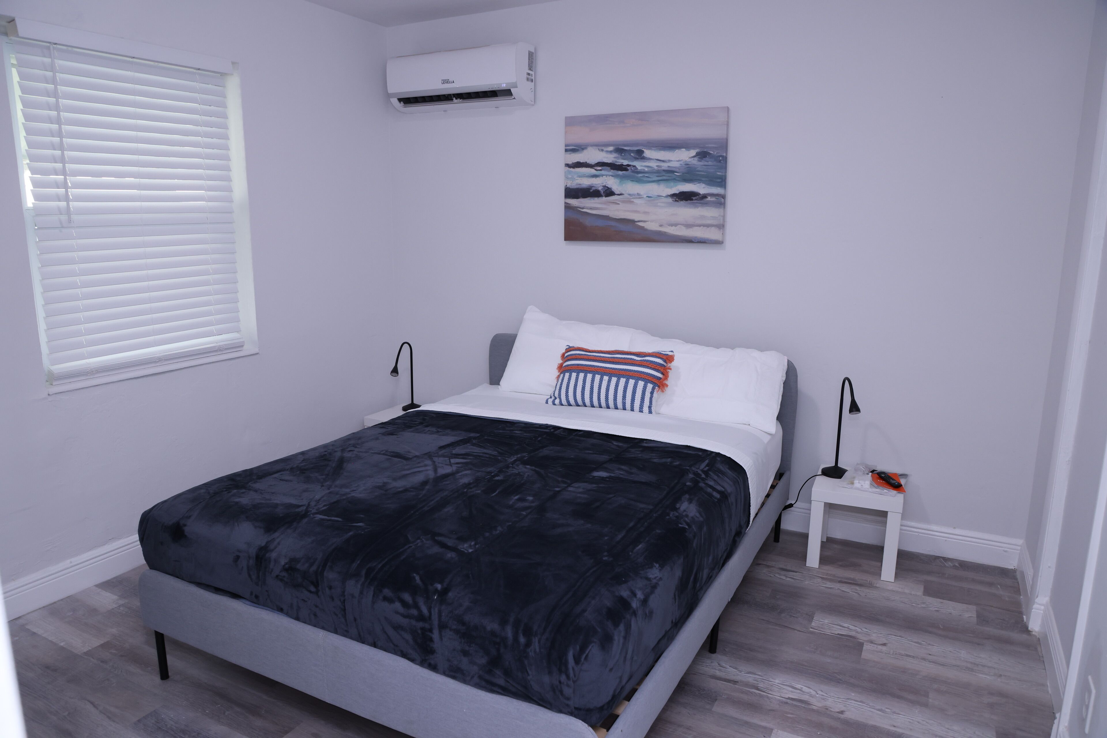 Quarto com cama, paredes brancas, pintura do oceano e janela com persianas.