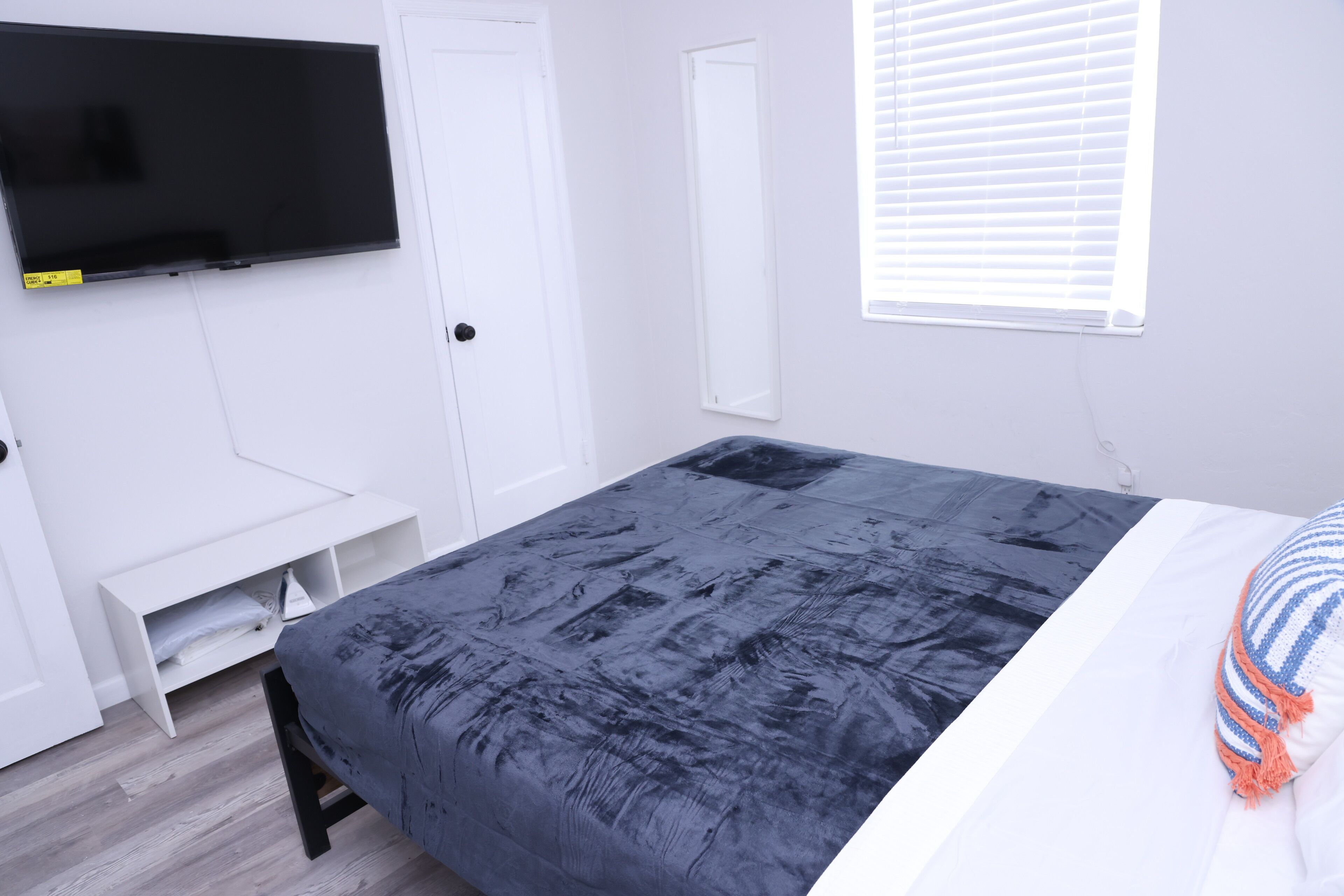 Ein Schlafzimmer mit einem Bett, einem an der Wand montierten Fernseher, einem Fenster mit Jalousien und einer weißen Tür.