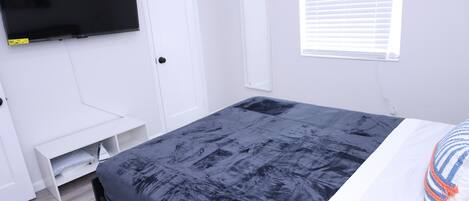 Ein Schlafzimmer mit einem Bett, einem an der Wand montierten Fernseher, einem Fenster mit Jalousien und einer weißen Tür.