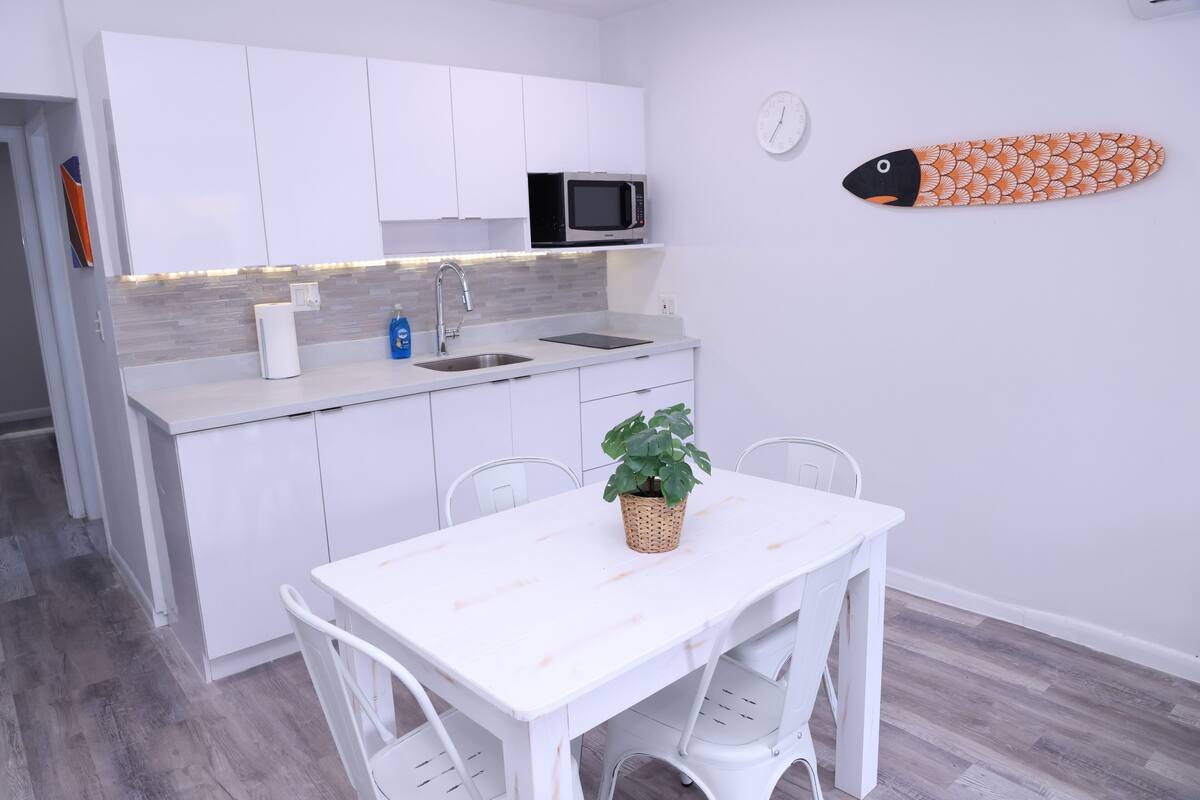 A compact kitchen with white cabinets, a microwave, and a sink. There's a white table with chairs and a potted plant.