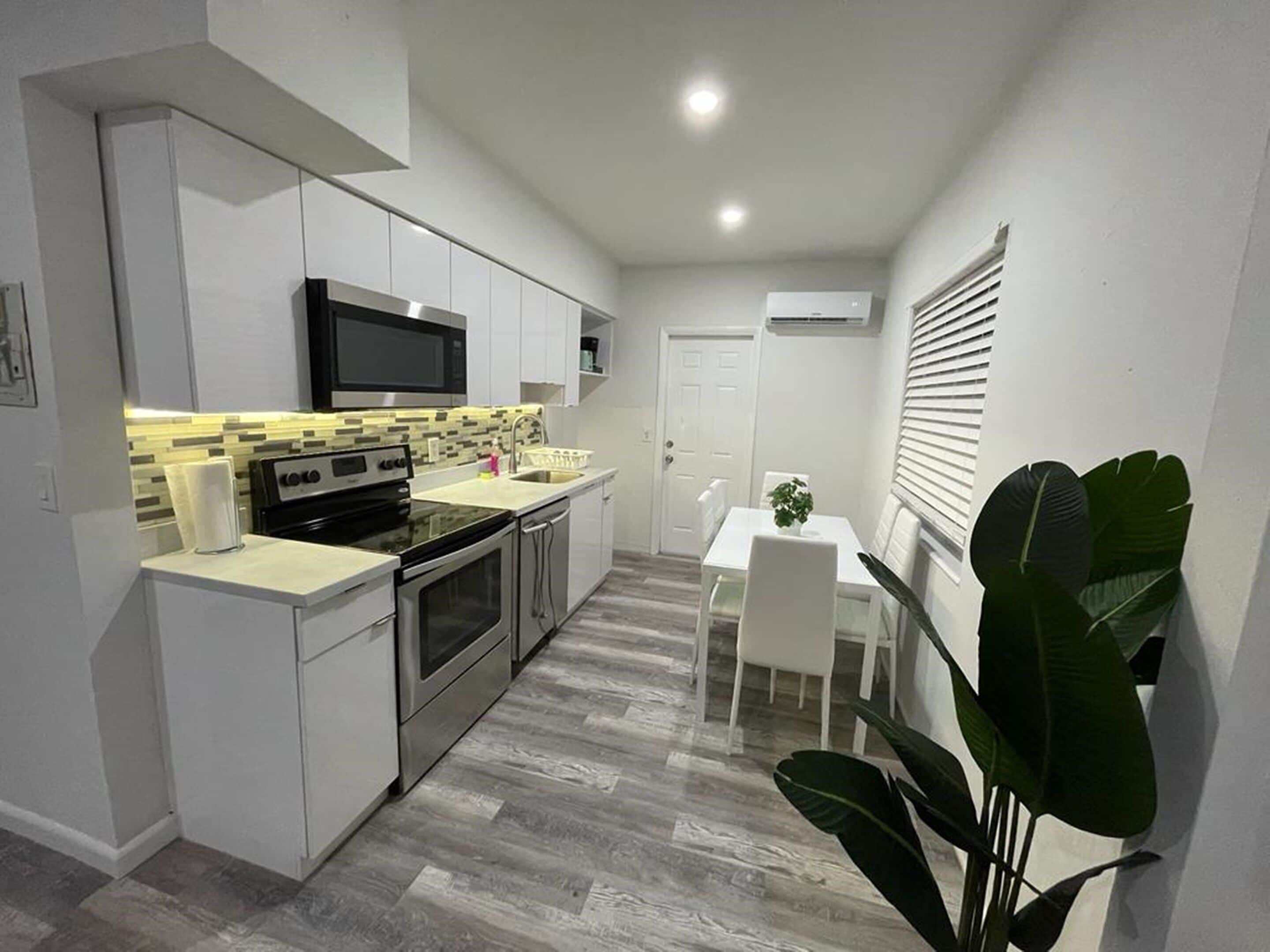 Une cuisine compacte avec des meubles blancs, un micro-ondes noir et un pare-feu en carreaux. On y trouve un coin repas avec une table et des chaises blanches, ainsi qu'une grande plante verte dans un coin.