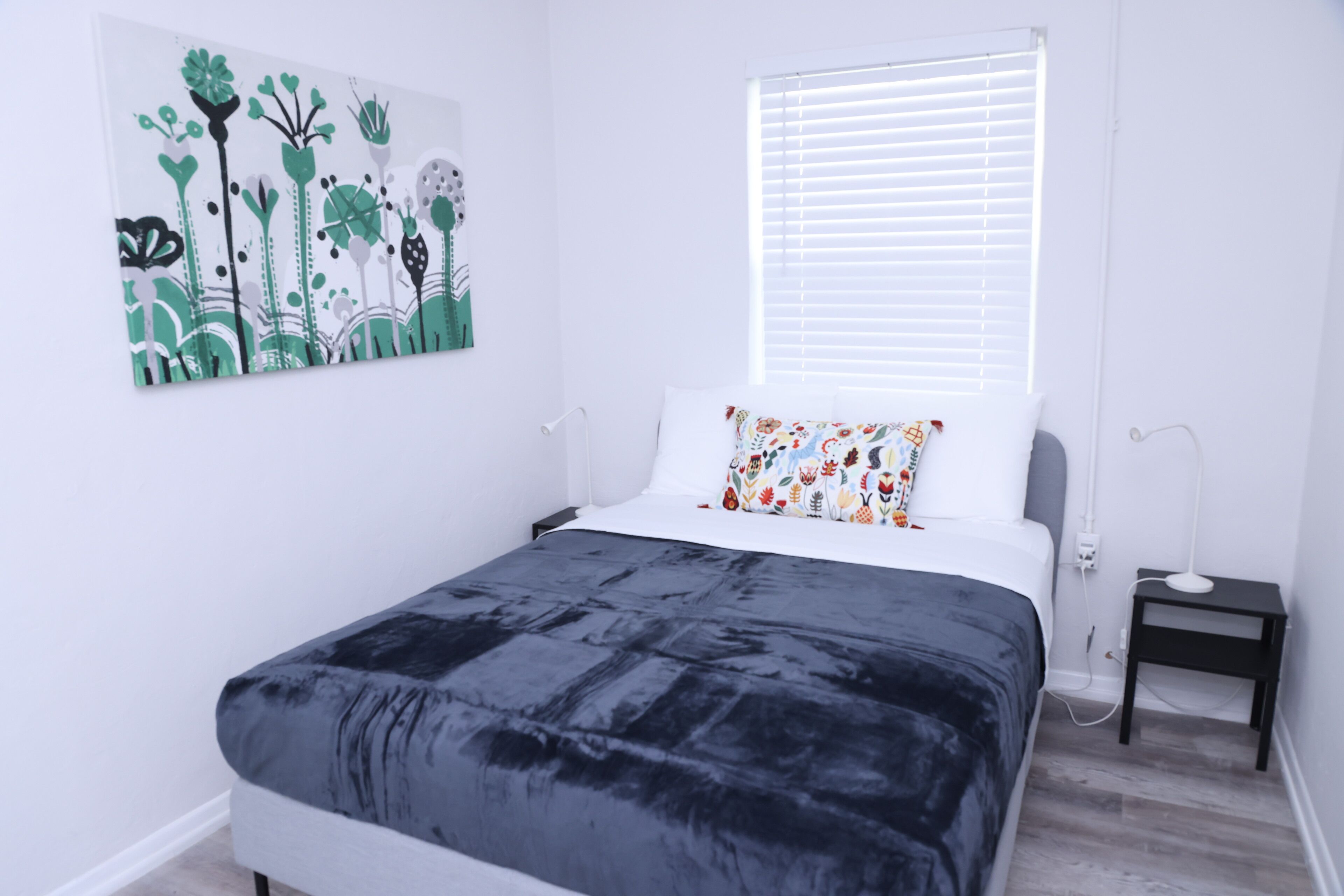 Ein Schlafzimmer mit Bett, Nachttisch, Lampe, einem Fenster mit Jalousien und einem Wandbild.