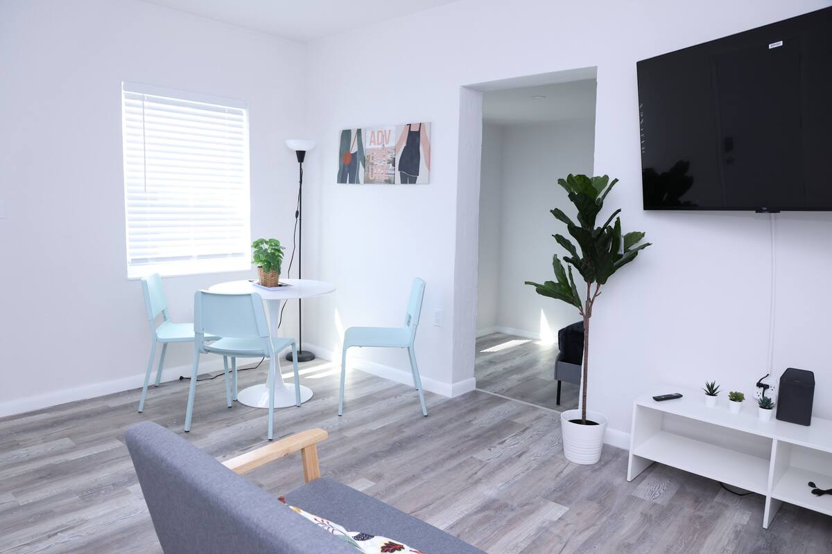 A modern living room with a grey sofa, a round dining table with two chairs, a large potted plant, a flat-screen TV mounted on the wall, and a white console table with small plants and a speaker.