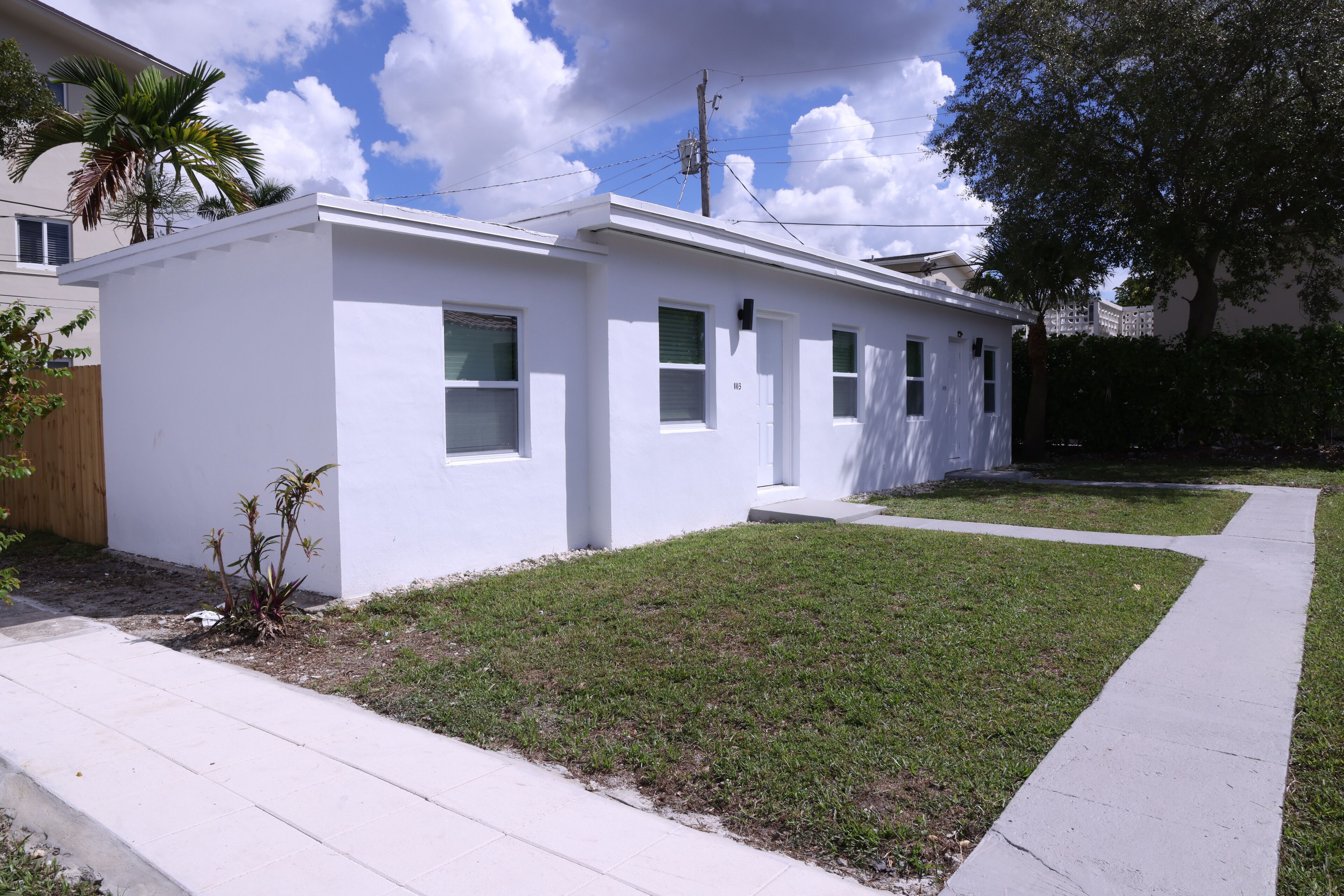 A modern single-story house with a flat roof, white exterior, and a small front yard with grass and a few plants.