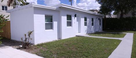 A modern single-story house with a flat roof, white exterior, and a small front yard with grass and a few plants.