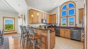 A spacious kitchen with wooden cabinets, a central island, and a large window offering a view of greenery. - Soundside Dog Friendly Home w/ Private Pool, Hot Tub, Elevator, and Game Room (Avon)